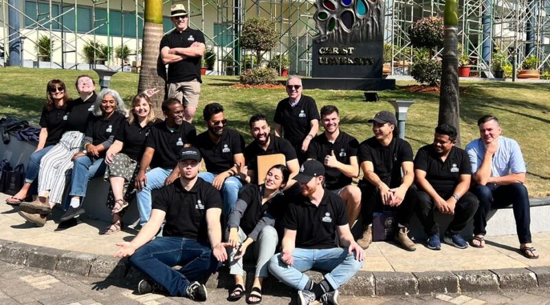 Team of 20+ youth in black t-shirts on lawn of building in India.