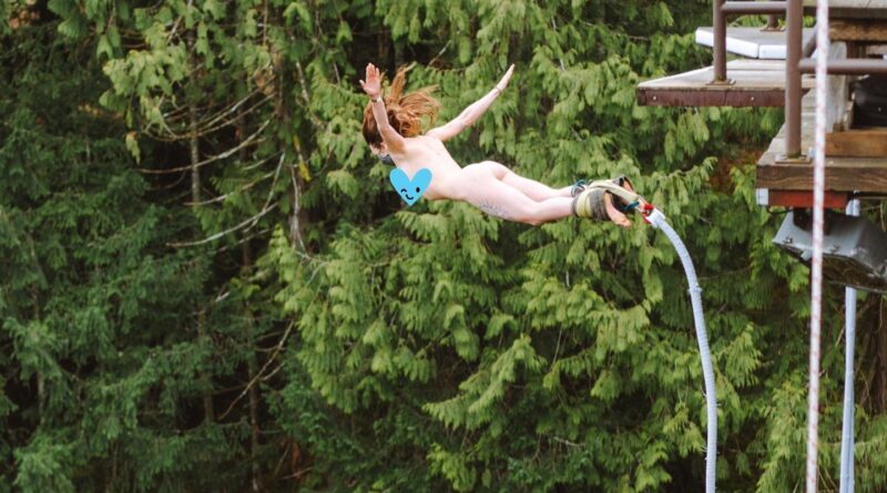 Woman mid-air bunny jumping naked against forest of trees.