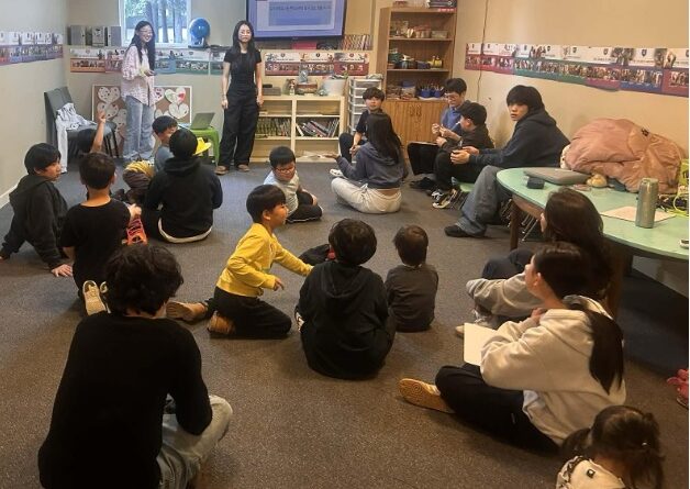 Korean children sitting on the floor in a classroom with two female instructors at the front of the room.