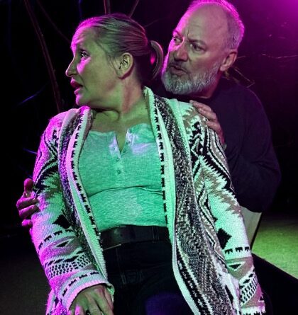 Man gripping womans arms from behind and looking at her as she looks away on stage