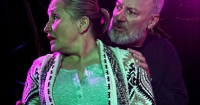 Man gripping womans arms from behind and looking at her as she looks away on stage