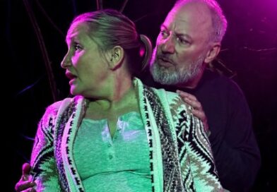 Man gripping womans arms from behind and looking at her as she looks away on stage